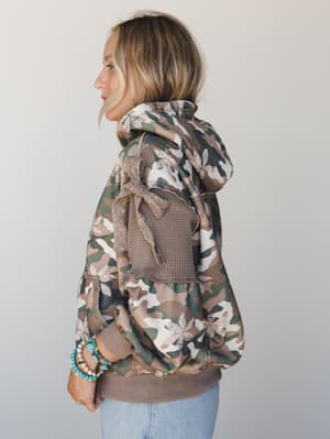 A side-profile shot of a camo hoodie jacket with unexpected boho charm — floral print mixed into the camo, a waffle-knit back panel, and a bow tie detail, styled with light wash jeans and stacked turquoise bracelets.