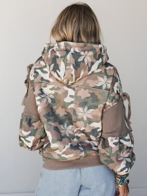 Shot from behind, a camo hoodie jacket with floral overlay print and waffle-knit sleeve panels does that rare thing — it's tough and pretty at the same time, styled casually with light wash jeans and stacked turquoise bracelets.