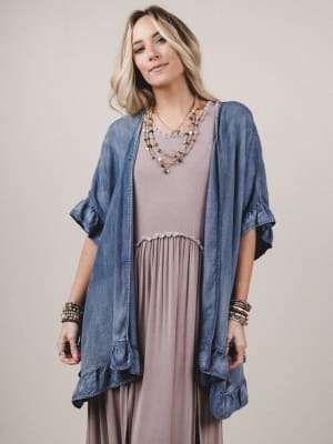 Woman wearing a denim cardigan over a long dress with layered necklaces on a plain background