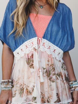A close-up of a denim kimono with a scalloped crochet hem tied at the center, layered over a coral embroidered top and a flowy floral skirt — the kind of outfit that looks like it came together effortlessly but definitely did not.