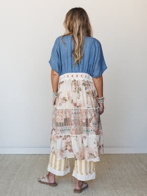 Shot from behind, a model wears a mixed-media kimono layered over wide-leg pants — the chambray top half transitions into a tiered, patchwork skirt with floral, geometric, and stripe panels connected by crochet trim, giving the whole look that effort