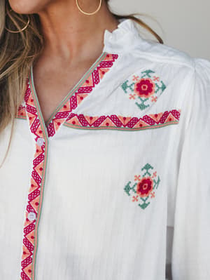 A close-up of a white button-front blouse with intricate multicolor embroidery along the placket and yoke — the kind of top that does all the talking so you don't have to.