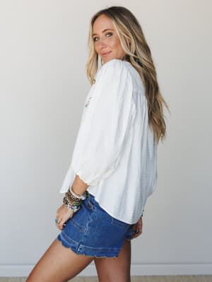 A breezy white embroidered blouse with billowy sleeves is styled with scallop-hem denim shorts and a stack of colorful beaded bracelets — the kind of effortless summer outfit that looks like it came together in five minutes but somehow feels complete