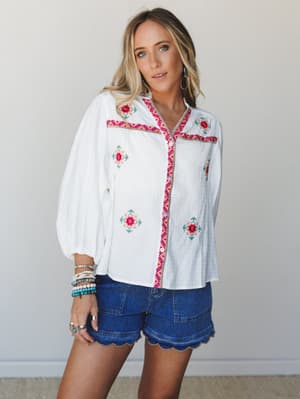 A breezy white embroidered blouse paired with scallop-hem denim shorts — the kind of outfit that looks equally at home on a farmers market Saturday or a summer trip somewhere with cobblestone streets.