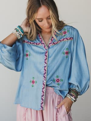 A soft chambray blouse with folk-inspired floral embroidery and contrast trim styling, paired with a blush skirt and stacked bracelets — the kind of outfit that looks like it came together effortlessly but definitely did not.