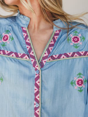 A close-up detail shot of a soft chambray blouse showing intricate floral and geometric embroidery across the yoke, with a zigzag-trimmed button placket and a delicate ruffle collar — the kind of top that looks like it has a whole story already.