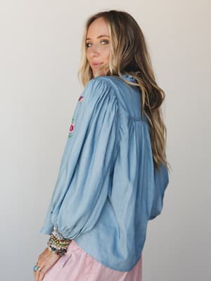 A soft washed denim blouse shot from a 3/4 back angle, its billowy balloon sleeves and floral embroidery giving it that lived-in, romantic feel — styled with a blush pink skirt and a stack of colorful bracelets that feel collected, not coordinated.