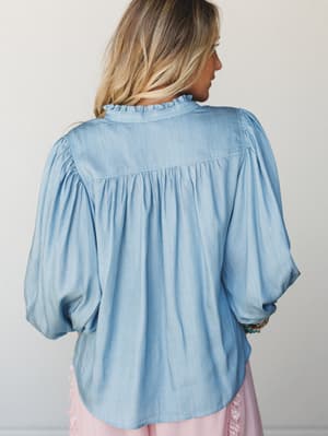 A soft washed denim blouse shot from the back, showing off its relaxed gathered silhouette, ruffle collar, and voluminous balloon sleeves — the kind of top that feels as good as it looks.