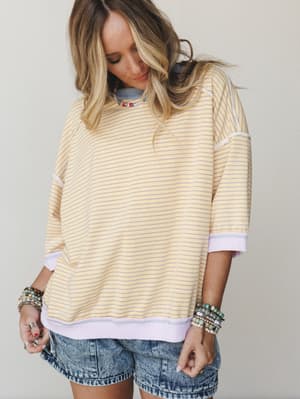 A relaxed striped sweatshirt top in buttery yellow and lavender is styled with acid wash denim shorts and stacked beaded bracelets — the kind of effortless warm-weather outfit that feels pulled-together without trying.