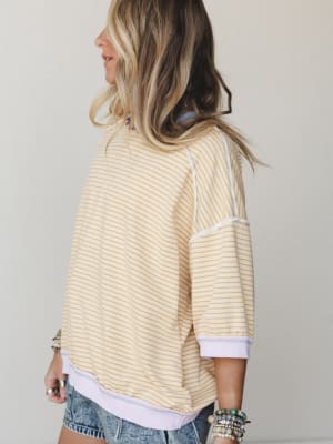 A relaxed striped pullover top in buttery yellow and lavender, shot from the back at a three-quarter angle — the exposed seam detailing and ribbed hem give it that lived-in, effortlessly cool feel, styled with cutoff denim shorts and layered beaded b
