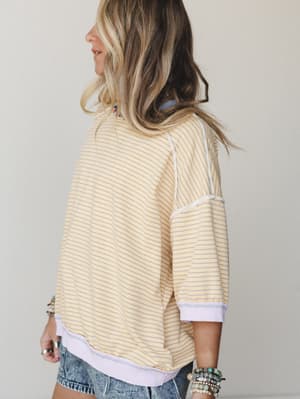 A relaxed striped pullover top in buttery yellow and lavender, shot from the back at a three-quarter angle — the exposed seam detailing and ribbed hem give it that lived-in, effortlessly cool feel, styled with cutoff denim shorts and layered beaded b