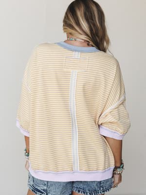 Shot from the back, this oversized striped pullover in buttery yellow and lavender is styled with acid wash denim shorts and layers of beaded bracelets — casual and a little nostalgic, like something you'd grab off a friend's chair and never give bac