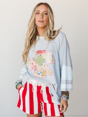 A breezy, oversized sweatshirt in soft cloud blue with a whimsical patchwork cross appliqué gets paired with bold red-and-white striped shorts and stacked bead bracelets — the kind of outfit that feels like a Saturday with nowhere to be and everywher