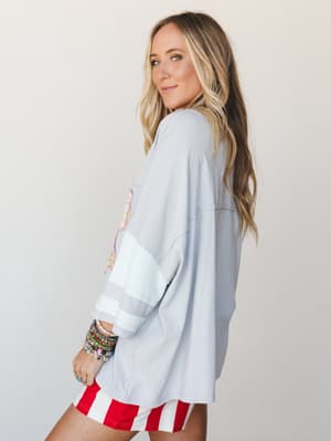 A side-profile shot of a model in an oversized cloud blue spirit jersey-style top with a colorful graphic and stripe sleeve detail, paired with bold red and white striped shorts and stacked beaded bracelets — casual and playful with a throwback-summe