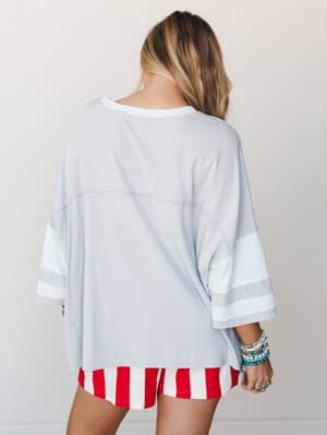 Shot from the back, a relaxed oversized jersey-style top in soft cloud blue is paired with bold red and white striped shorts and a stack of colorful beaded bracelets — casual, a little retro, and completely unpretentious.