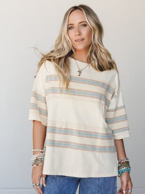 A relaxed, oversized striped tee in creamy white with dusty blue and mauve stripes, styled casually with denim and stacked boho bracelets — the kind of top that looks effortless whether you grabbed it intentionally or just reached for the first thing