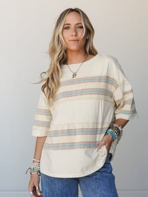 A relaxed, oversized striped tee in a soft cream base with dusty blue and terra cotta stripes, styled with wide-leg jeans and stacked beaded bracelets — the kind of effortless top that makes a Tuesday feel a little more intentional.