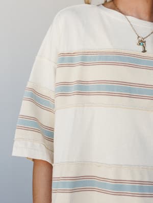 A close-up of a relaxed striped tee in creamy off-white with soft blue and dusty rose horizontal stripes, styled with a layered charm necklace — the kind of top that looks like it was made for slow mornings and beach-town errands.