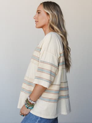 A side-profile shot of a relaxed, oversized striped tee in creamy ivory with dusty blue and terracotta stripes — styled with a stack of beaded bracelets and jeans for that effortlessly lived-in feel.