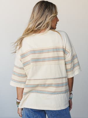 Shot from the back, a relaxed oversized tee in creamy banana milk with soft sage and terracotta horizontal stripes — styled with denim and stacked beaded bracelets for that effortless, lived-in weekend feel.