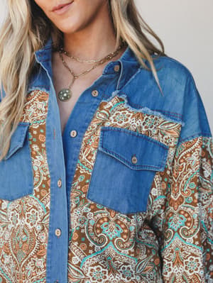 A mixed-fabric button-up shacket combining medium-wash denim panels with a warm brown and turquoise paisley print — styled with layered gold chain necklaces for that effortless Western-boho feel.