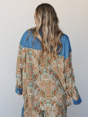 A back-facing shot of a relaxed paisley top with denim yoke and cuffs, the warm brown-and-turquoise print doing all the talking while the chambray detail grounds it — the kind of piece that feels like it belongs in a sunlit vintage shop.