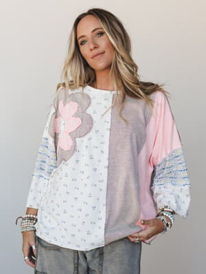A mixed-print oversized top in ivory and pink with a bold flower appliqué, styled with stacked bracelets and gray jeans — the kind of top that does all the talking so you don't have to.