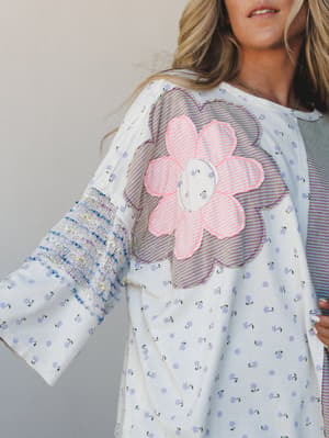 A close-up of a relaxed mixed-print top in ivory with a ditsy lavender floral print, featuring a bold patchwork flower appliqué in pink and purple stripes at the chest — the kind of piece that feels like it was made from your favorite vintage scraps.