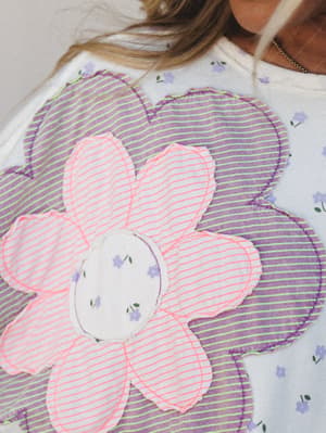A close-up of an ivory top covered in a small floral print, featuring oversized patchwork flower appliqués in striped pink and purple fabric with contrast neon stitching — the kind of piece that makes you smile before you've even put it on.