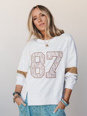 A relaxed varsity-style sweatshirt in heathered white with a floral-print '87' graphic and tan sleeve stripes, styled with floral blue pants and stacked boho bracelets — casual nostalgia with a soft, feminine twist.