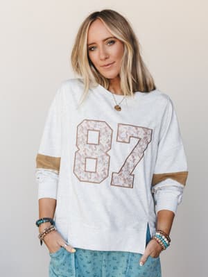 A relaxed oversized sweatshirt in light heather gray with a floral-appliqué varsity number graphic and tan sleeve stripe detail, styled with stacked boho bracelets and a peek of blue floral pants — the kind of top that makes a Saturday feel intention