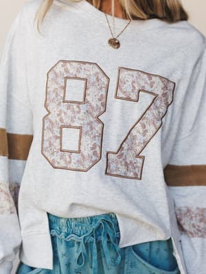 A close-up shot of a heathered gray sweatshirt with a floral-appliqué '87' varsity graphic and contrast stripe sleeves, styled with a dusty blue drawstring skirt and a delicate gold charm necklace — casual-cool with a soft, nostalgic edge.