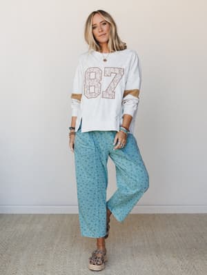 A relaxed, mix-and-match outfit built around a heather gray varsity sweatshirt with a floral-filled '87' graphic and gold color-block sleeves, paired with wide-leg teal floral pants and espadrille sandals — the kind of effortless combo that looks lik