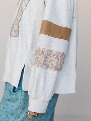 A close-up detail shot of a heather gray sweatshirt-style top with mixed-fabric patchwork sleeves — suede-like tan and dusty floral print panels layered against soft knit, styled with light blue printed denim and silver stacked rings.