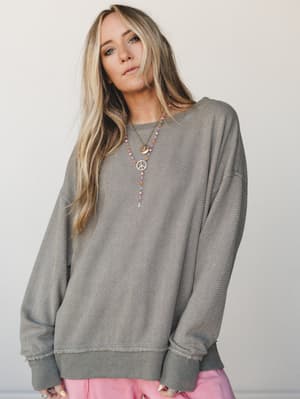 An oversized textured sweatshirt in muted olive worn with layered boho necklaces and a peek of pink pants — the kind of effortless, lived-in look that feels intentional without trying.