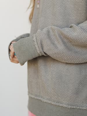 A close-up detail shot of a textured olive sweatshirt with raw-edge finishing and ribbed cuffs — the kind of top that looks like it's been lived in for years in the best possible way.