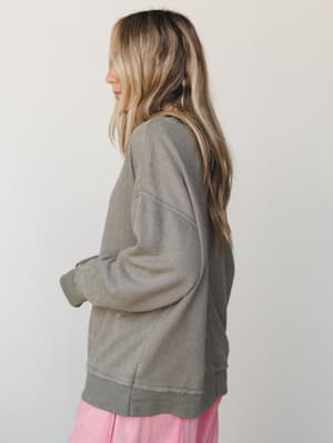 A side-profile shot of a model in an oversized olive textured sweatshirt with visible seam detailing, paired with a soft pink skirt — the kind of effortless combo that looks accidental but isn't.