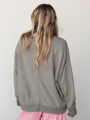 Shot from the back, a model wears an oversized textured knit pullover in washed olive with dropped shoulders and ribbed hem, paired with soft pink wide-leg pants — the kind of effortless layering that looks like it happened by accident and somehow wo