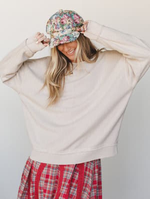 A smiling model adjusts a floral baseball cap while wearing an oversized cream textured sweatshirt layered over a red plaid midi skirt — the kind of effortlessly mixed outfit that looks like it came together on a really good morning.