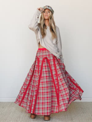 A full-body model shot of a flowy red plaid maxi skirt styled with an oversized oatmeal knit top, floral bucket hat, and brown clogs — the kind of mix-and-match outfit that looks effortless but clearly wasn't an accident.