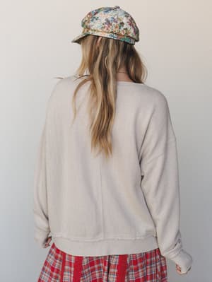 Shot from the back, she's wearing a relaxed textured sweatshirt in a warm mushroom tone layered over a red plaid midi skirt, topped off with a floral tapestry cap — the kind of mix-and-match that looks accidental in the best way.