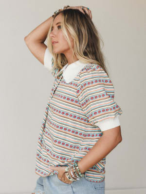 A side-profile studio shot of a striped floral top with a white peter pan collar, styled with stacked beaded bracelets and light wash denim — the kind of top that looks like it came from your most well-traveled friend's closet.