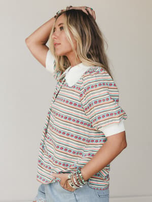 A side-profile studio shot of a striped floral top with a white peter pan collar, styled with stacked beaded bracelets and light wash denim — the kind of top that looks like it came from your most well-traveled friend's closet.