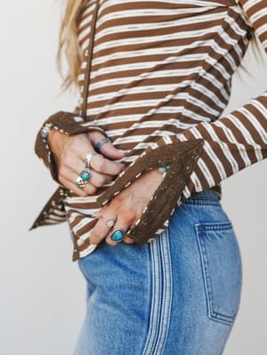 A close-up detail shot of a brown and cream striped long-sleeve top with lace-trimmed bell cuffs, styled with light wash denim and layered silver turquoise rings — the kind of outfit that looks like it was put together effortlessly but very much wasn