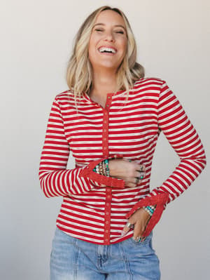 A laughing blonde model in a fitted red and cream striped henley top with lace cuff trim and snap buttons, paired with light wash jeans — the kind of top that makes a Tuesday feel a little more intentional.