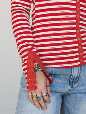 A close-up detail shot of a red and white striped long-sleeve top with lace cuff trim and snap button placket, styled with light-wash denim and layered boho jewelry — rings, turquoise beads, and stacked bracelets that feel very much like her everyday