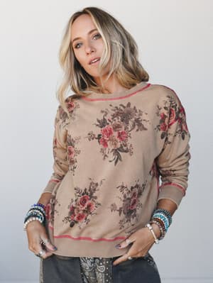 A relaxed floral sweatshirt in warm toast with vintage rose clusters and contrast stitching — styled with stacked beaded bracelets and a peek of paisley beneath, the whole look has that effortlessly collected, lived-in feeling.