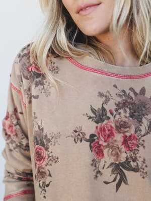 A close-up detail shot of a toasty tan sweatshirt with moody vintage floral bouquets and a pop of pink contrast stitching at the neckline — the kind of top that feels like a hug and still looks intentional.