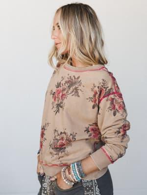 A floral sweatshirt in warm tan worn three-quarter back by a blonde model stacked in beaded bracelets — the kind of top that looks like it's been loved for years and somehow gets better every wash.