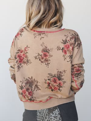 A back-view shot of a floral printed sweatshirt in warm toast, scattered with vintage rose bouquets and finished with red contrast stitching at the hem and seams — the kind of top that feels like a hug with a little something extra.
